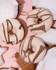 This image shows three of the same donut-shaped bath bombs arranged among open coconut halves on a pastel pink background. The bath bombs retain their off-white base with brown decorative swirls, visually reinforcing the "Coconut Vanilla" theme with a tropical and indulgent aesthetic.