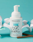 white bottle with white pump. background is teal. next to it are paper flowers and vanilla sticks