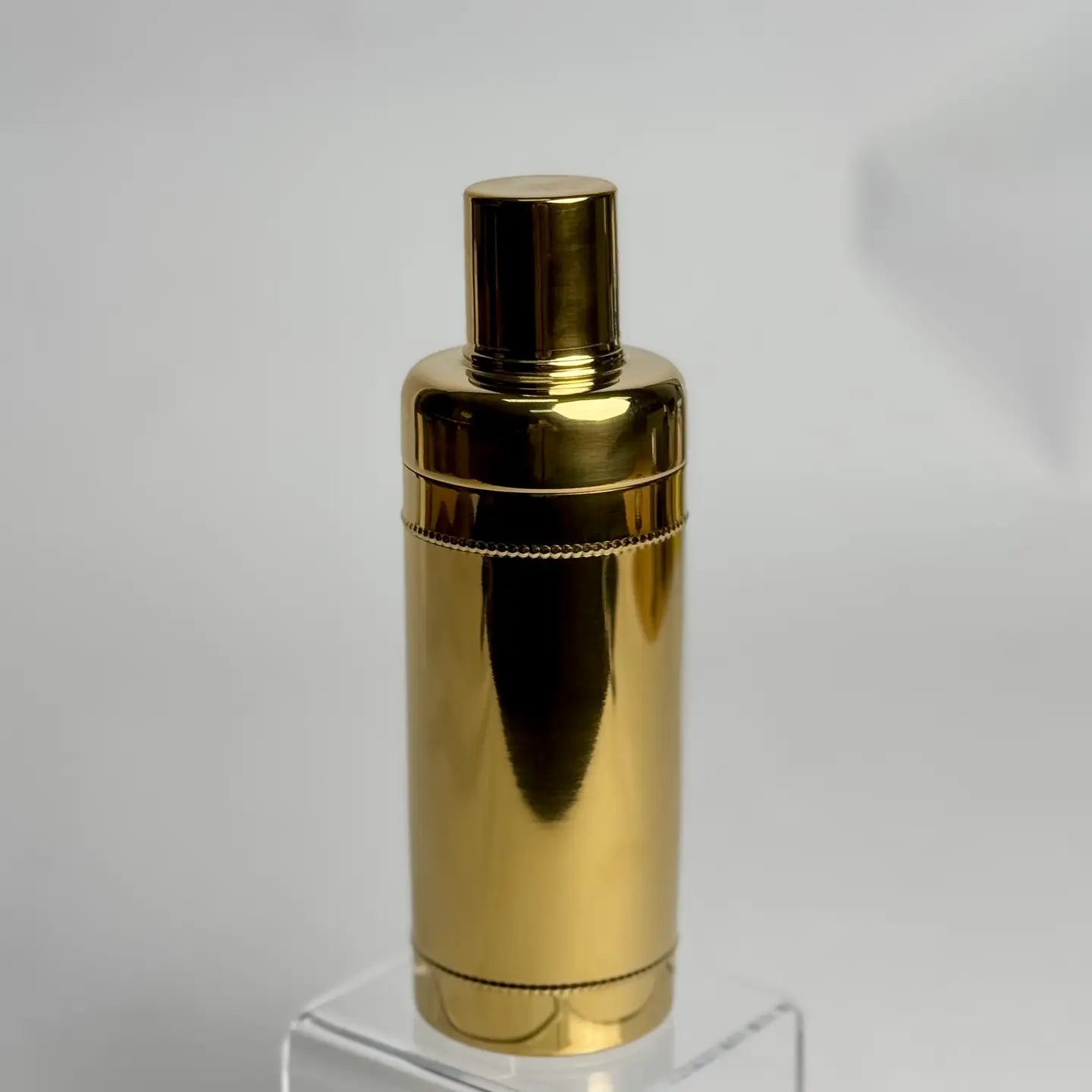 Gold metallic cocktail shaker on a clear stand with a white background