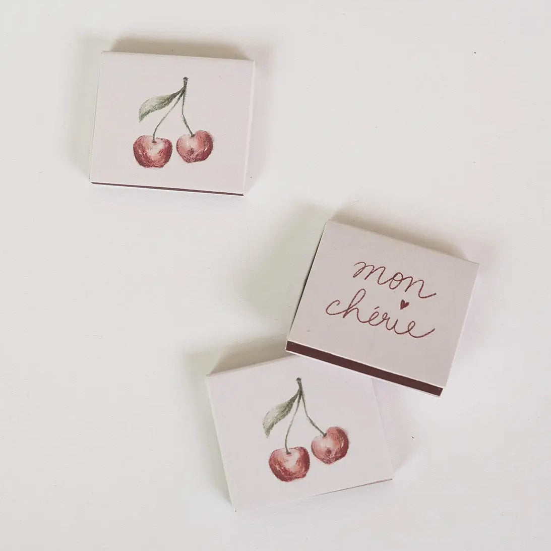Three small rectangular boxes arranged on a white background. Two boxes feature a watercolor-style red cherry illustration, and one box has the words 'mon chârie' in metallic rose-gold script.