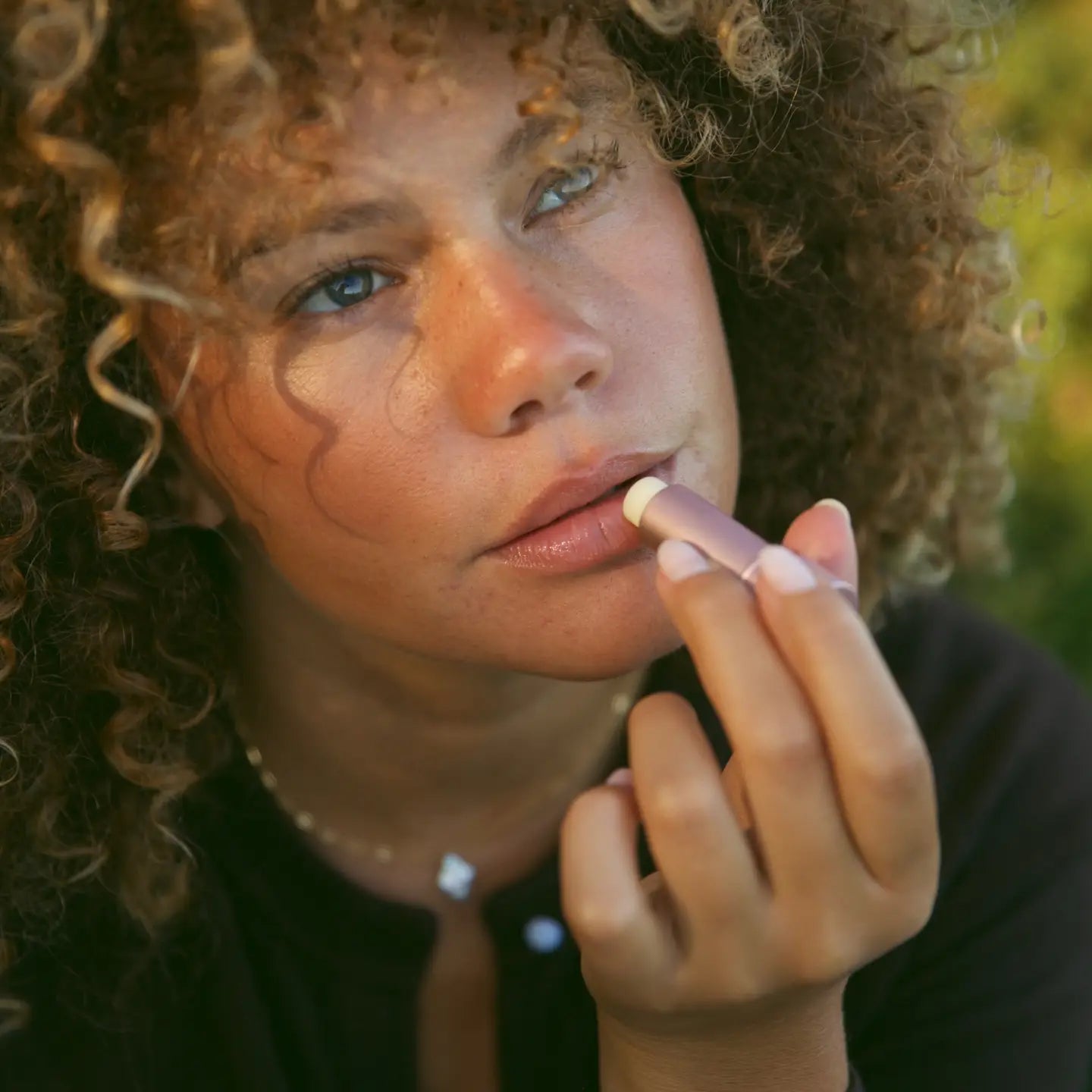 Woman applying lipstick outdoors with a blurred natural background