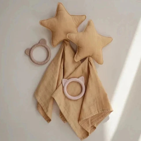 Two yellow star-shaped plush lovey blankets with attached muslin fabric are laid out on a soft beige surface. Two circular teething rings with bear ears—one in light brown and the other in pink—are placed next to the blankets, creating a cozy and playful arrangement.