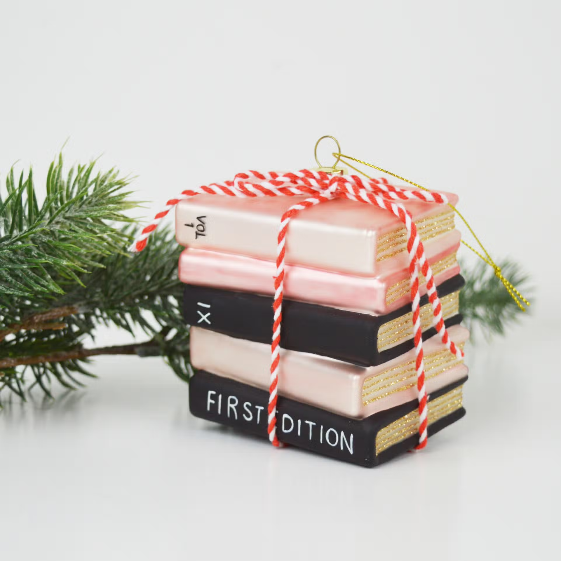 ornament shaped like a stack of book tied together with red and white twine. Book are pink and black with gold glitter showing the pages. 