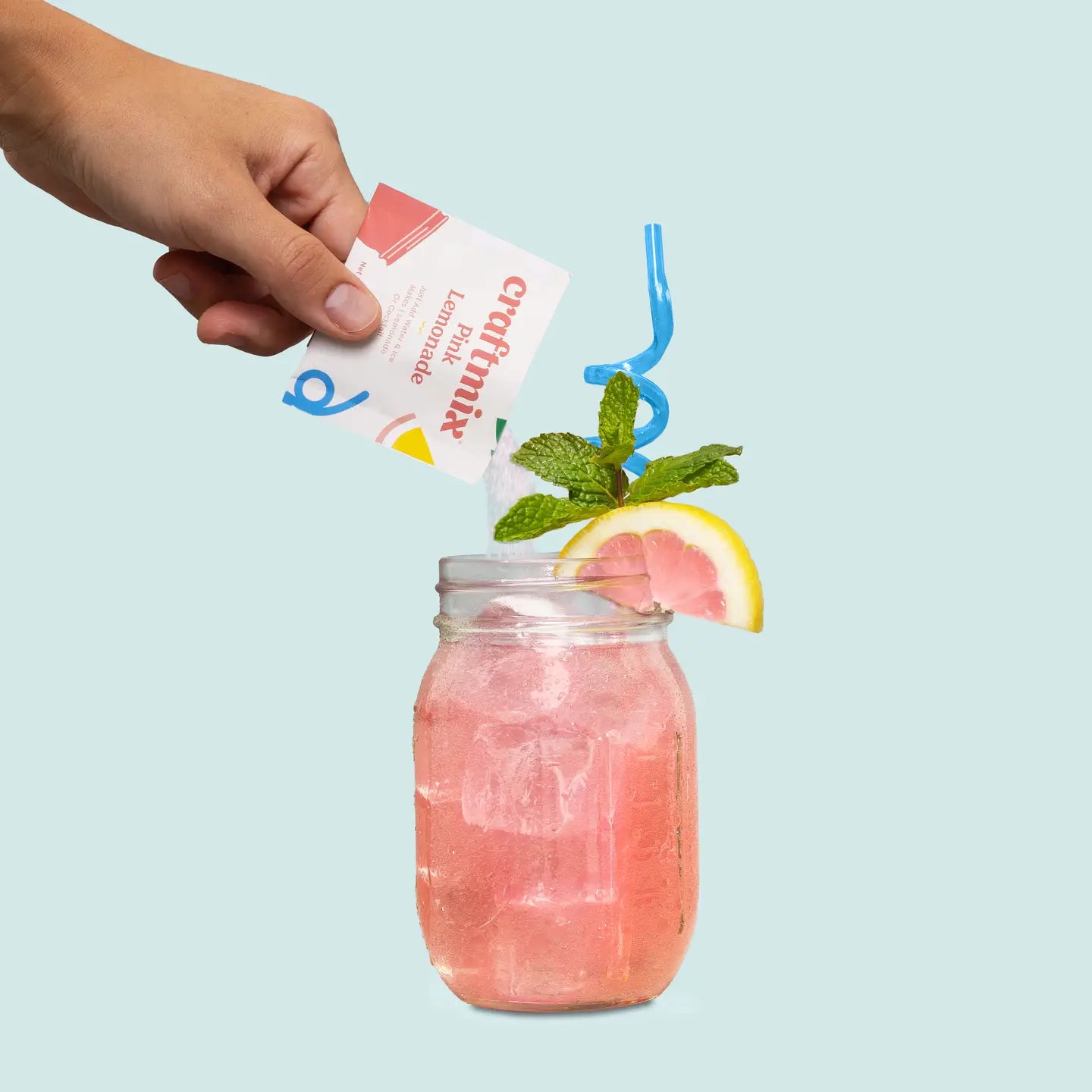 Hand holding a packet of 'craftymix' next to a pink cocktail with a lemon wedge and mint leaves on a light blue background