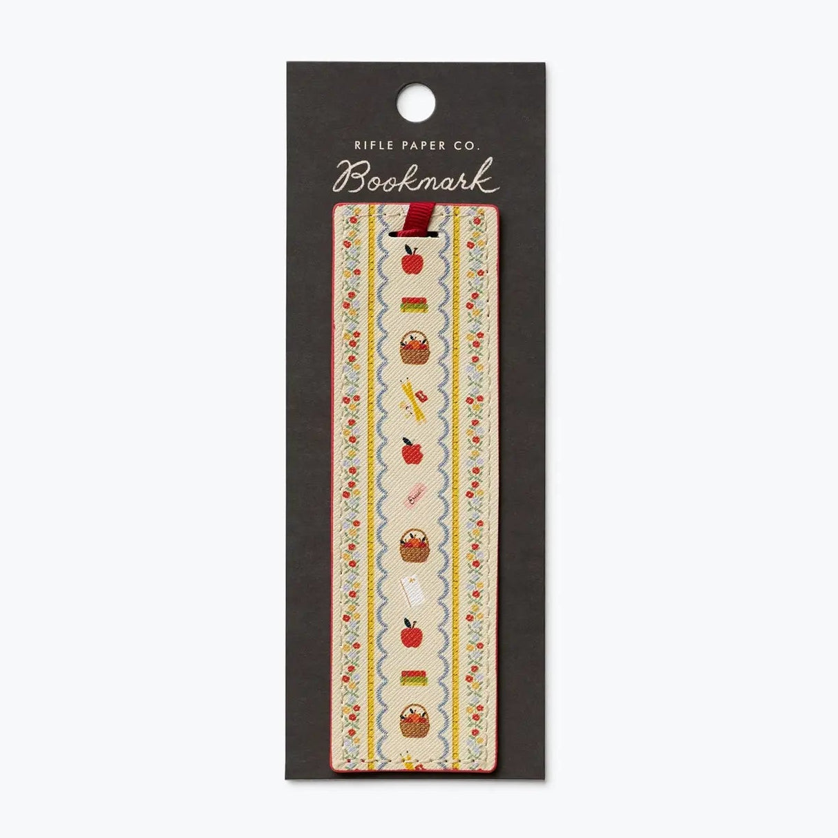A decorative bookmark from Rifle Paper Co. in its original black hanging card packaging, featuring a vertical strip with a cream background and a whimsical pattern of red apples, brown baskets, and small yellow flowers.