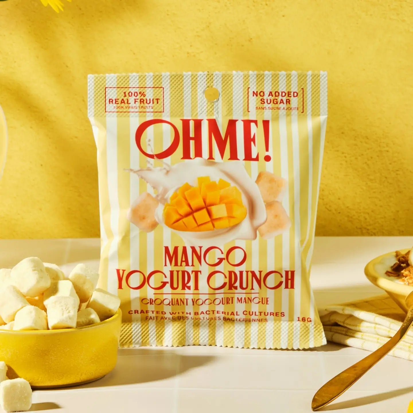 A bag of Ohme! Mango Yogurt Crunch is displayed on a white surface with a yellow background. The packaging features a cream and yellow vertical stripe pattern with a central graphic of a sliced mango in yogurt.