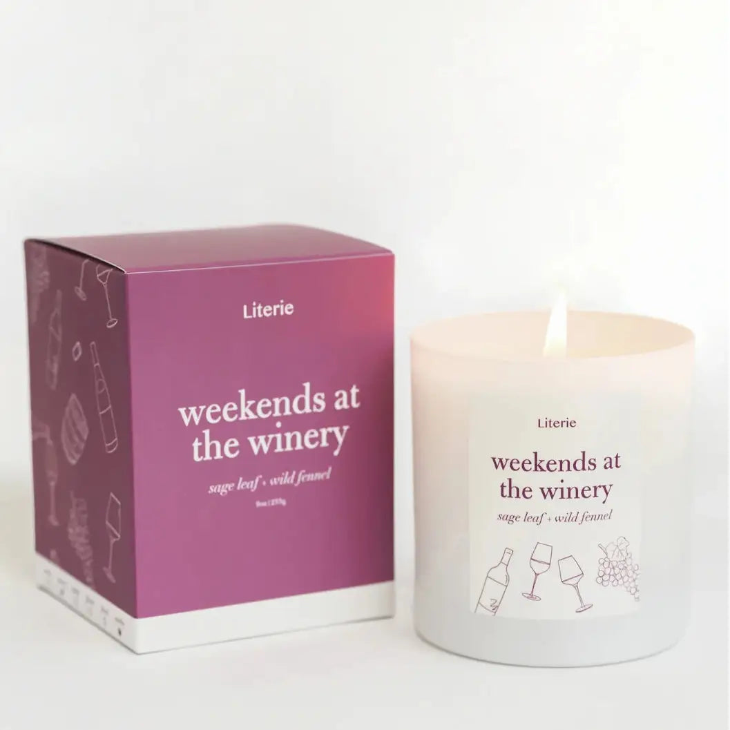 A lit white scented candle next to its magenta and white packaging box, both labeled 'Literie weekends at the winery' with wine-themed line drawings.