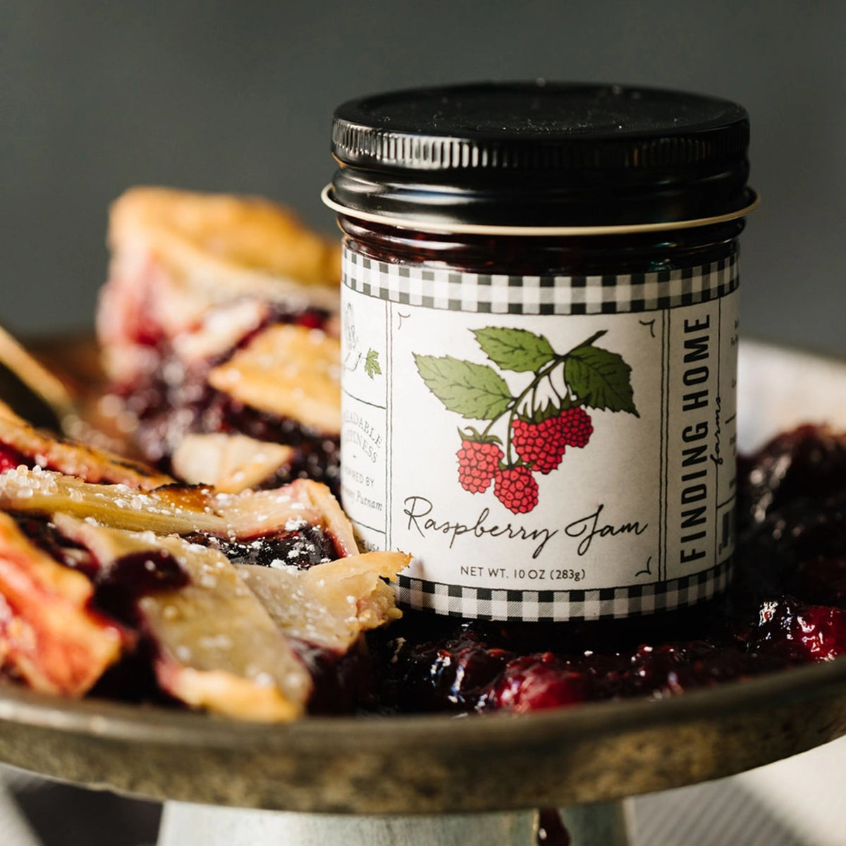 Jar of raspberry jam labeled 'Finding Home' on a plate with pie slices.