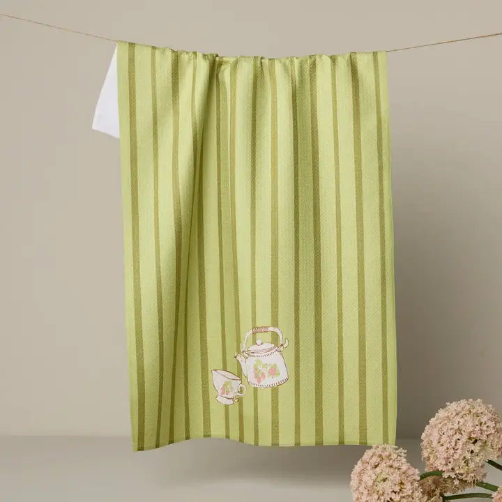 Green striped towel with teapot and cup design hanging on a line against a beige wall.