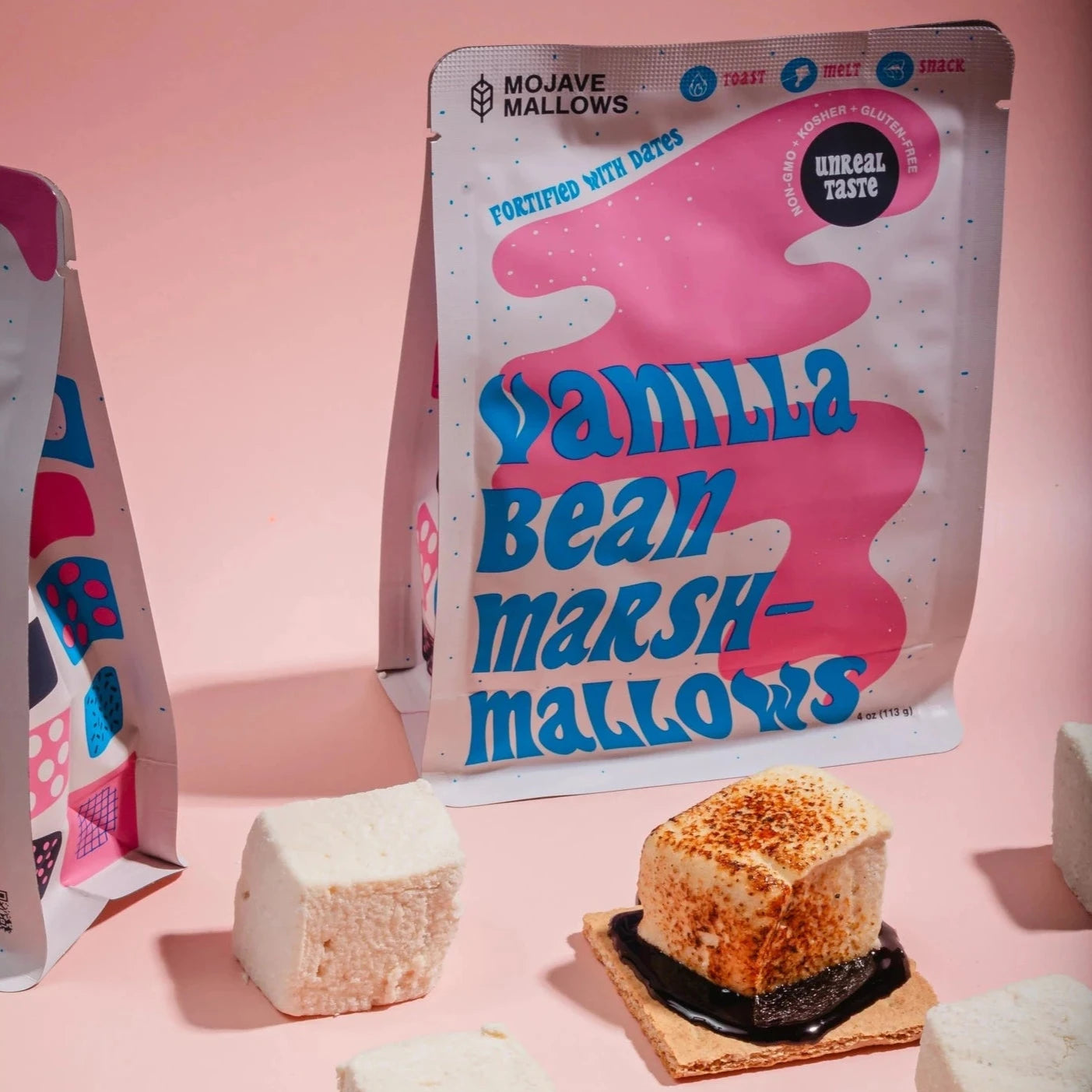 A package of Mojave Mallows Vanilla Bean Marshmallows is showcased on a pastel pink background. Large, fluffy white marshmallows are scattered around the package, with one toasted marshmallow sitting on a graham cracker with melted chocolate beneath it, creating a partial s'more.