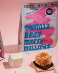 A package of Mojave Mallows Vanilla Bean Marshmallows is showcased on a pastel pink background. Large, fluffy white marshmallows are scattered around the package, with one toasted marshmallow sitting on a graham cracker with melted chocolate beneath it, creating a partial s'more.