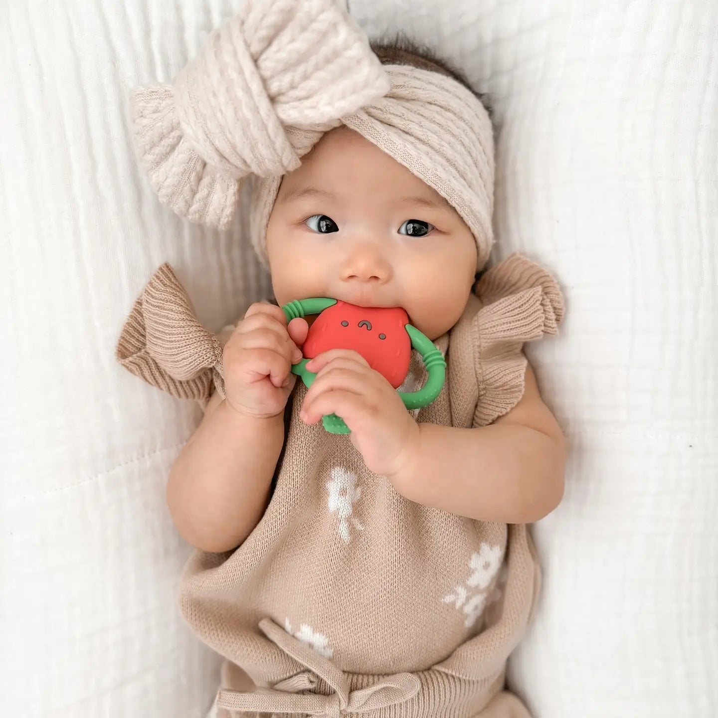 Baby wearing a beige outfit and headband, holding a red teething toy on a white background