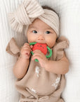 Baby wearing a beige outfit and headband, holding a red teething toy on a white background