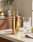 Gold cocktail shaker on a kitchen counter with a glass of ice and lemons.