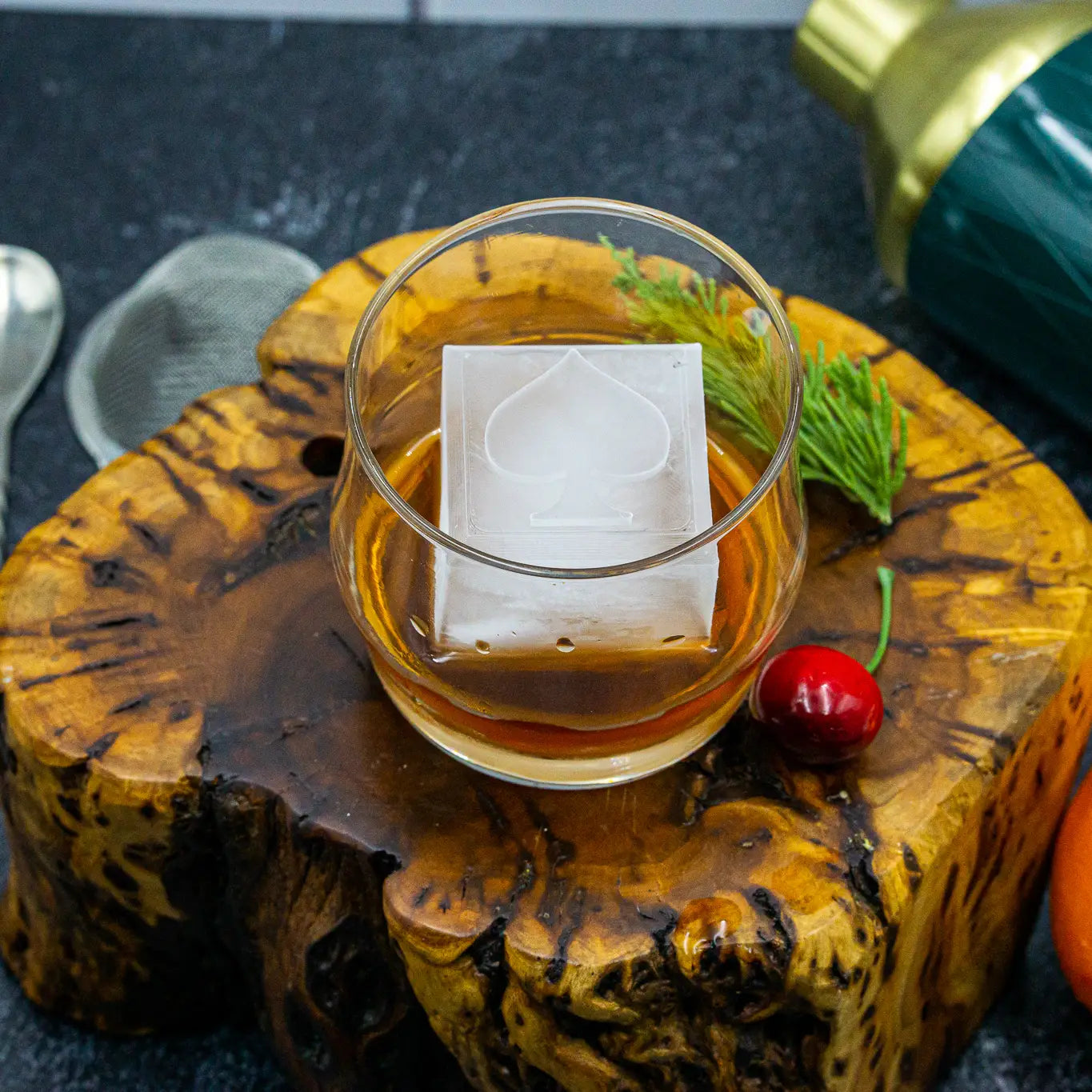 Glass of amber liquid with a decorative ice cube on a wooden coaster, surrounded by a cherry and greenery.