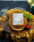 Glass of amber liquid with a decorative ice cube on a wooden coaster, surrounded by a cherry and greenery.