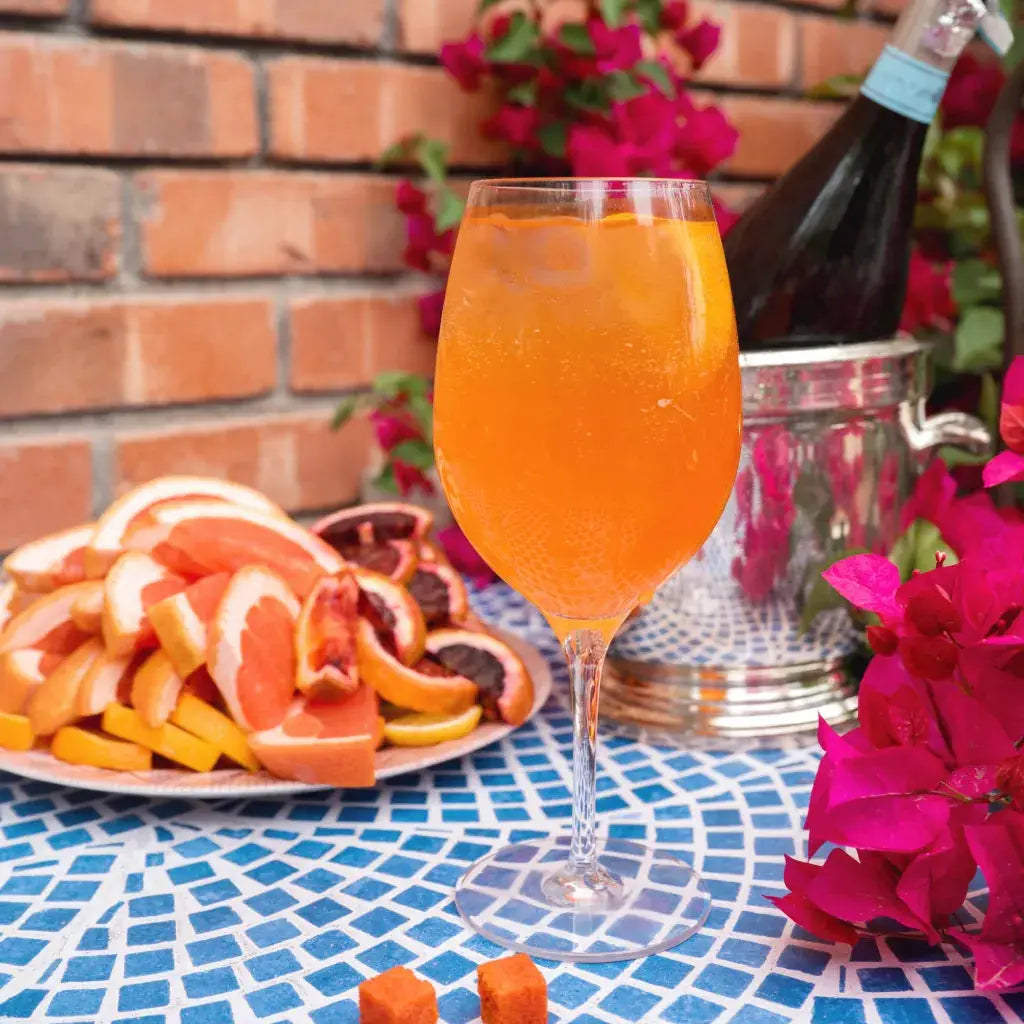 An orange-colored sparkling beverage in a wine glass, set on a blue mosaic tablecloth next to a plate of sliced citrus fruits, a bottle in an ice bucket, and bright pink flowers against a brick wall background.