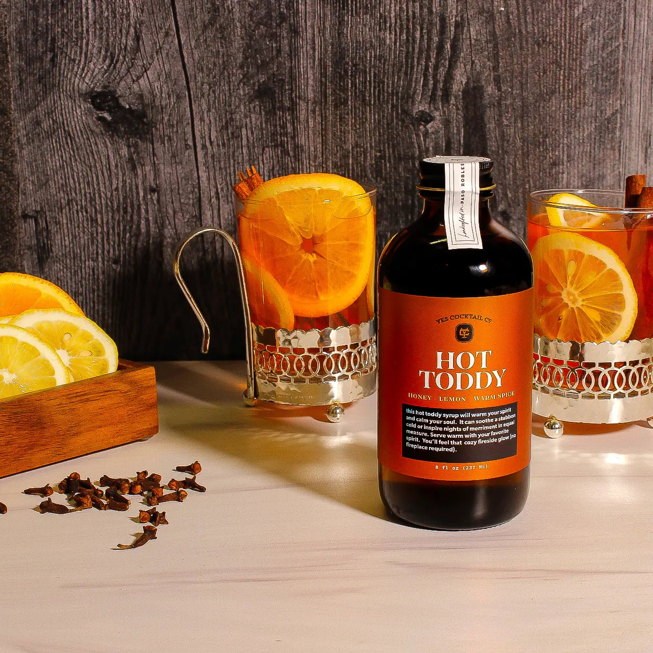 Hot Toddy bottle with glasses of hot toddy garnished with oranges on a wooden surface.