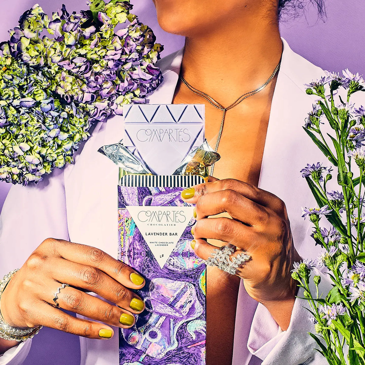 A person in a white blazer holds a lavender bar package from Compacts de Parfums, surrounded by purple and green flowers against a lavender background.
