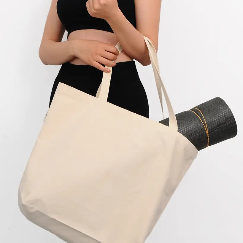 Person holding a beige tote bag with a black yoga mat underneath on a white background