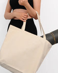 Person holding a beige tote bag with a black yoga mat underneath on a white background