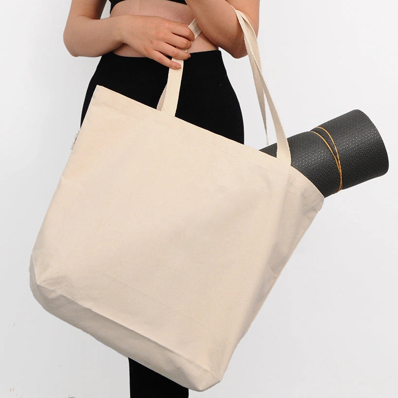 Person holding a beige tote bag with a yoga mat over their shoulder on a white background