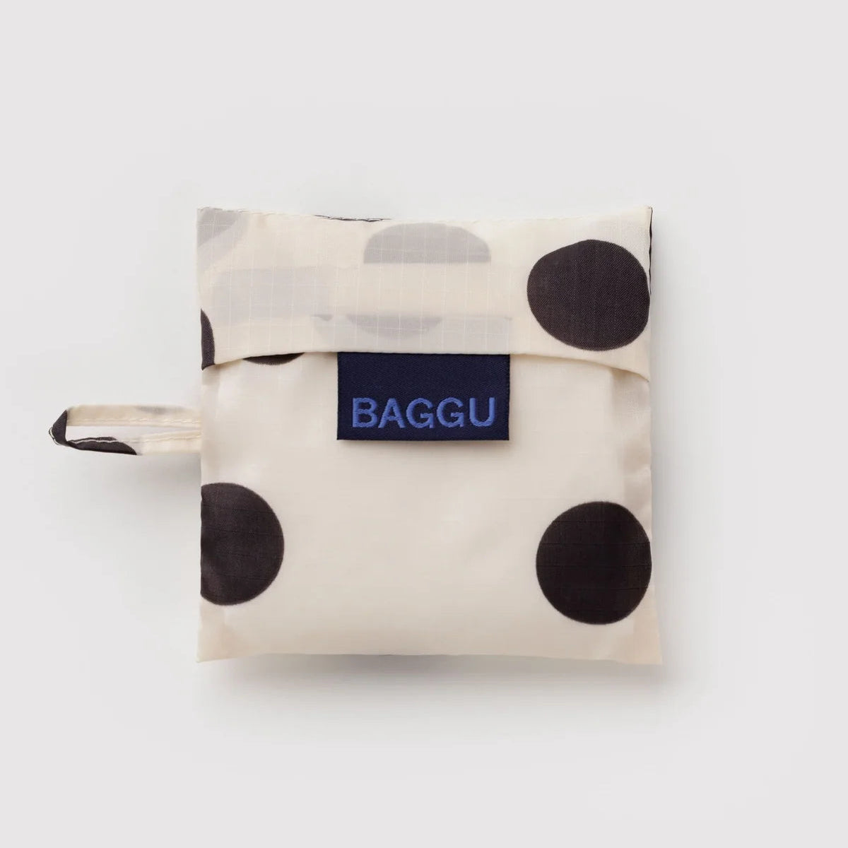 A folded reusable bag from BAGGU with a cream base, large black polka dots, and a navy blue brand label on the front.