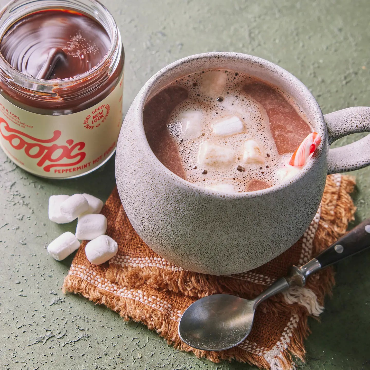 Ceramic mug with hot chocolate and marshmallows next to a jar of peppermint hot chocolate mix on a textured surface.