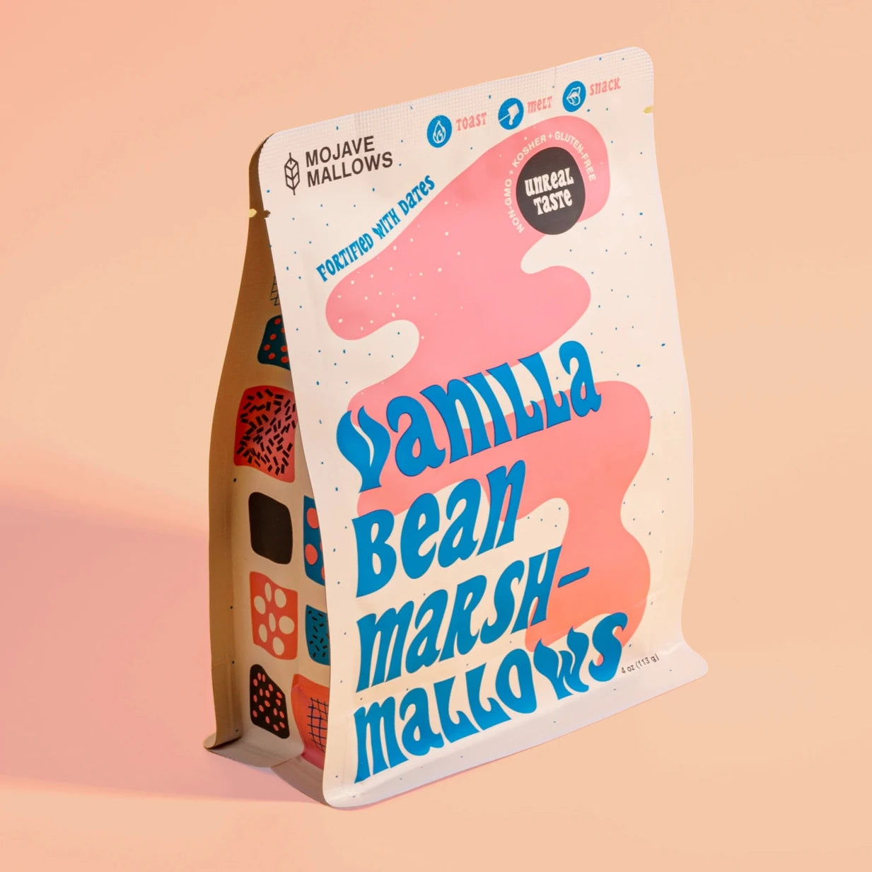 A standalone package of Mojave Mallows Vanilla Bean Marshmallows is photographed against a warm peach background. The bag's playful design features bold blue text and abstract pink and white patterns, highlighting that the marshmallows are fortified with dates and suitable for toasting, melting, or snacking.