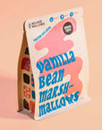 A standalone package of Mojave Mallows Vanilla Bean Marshmallows is photographed against a warm peach background. The bag's playful design features bold blue text and abstract pink and white patterns, highlighting that the marshmallows are fortified with dates and suitable for toasting, melting, or snacking.