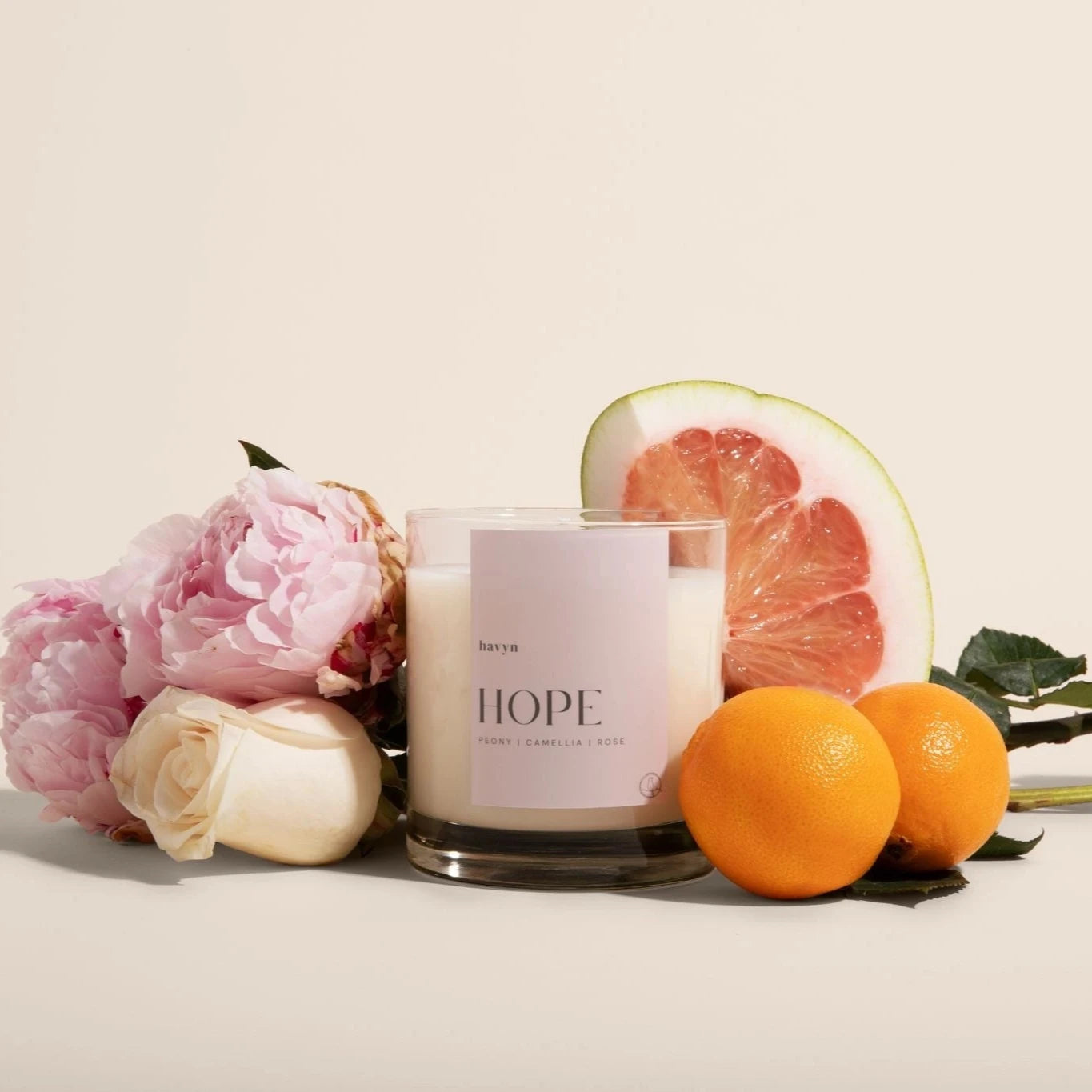 The second image displays the same candle surrounded by complementary elements such as flowers (peonies, roses) and citrus fruits like grapefruit and oranges, hinting at the fragrance profile of the product