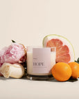 The second image displays the same candle surrounded by complementary elements such as flowers (peonies, roses) and citrus fruits like grapefruit and oranges, hinting at the fragrance profile of the product