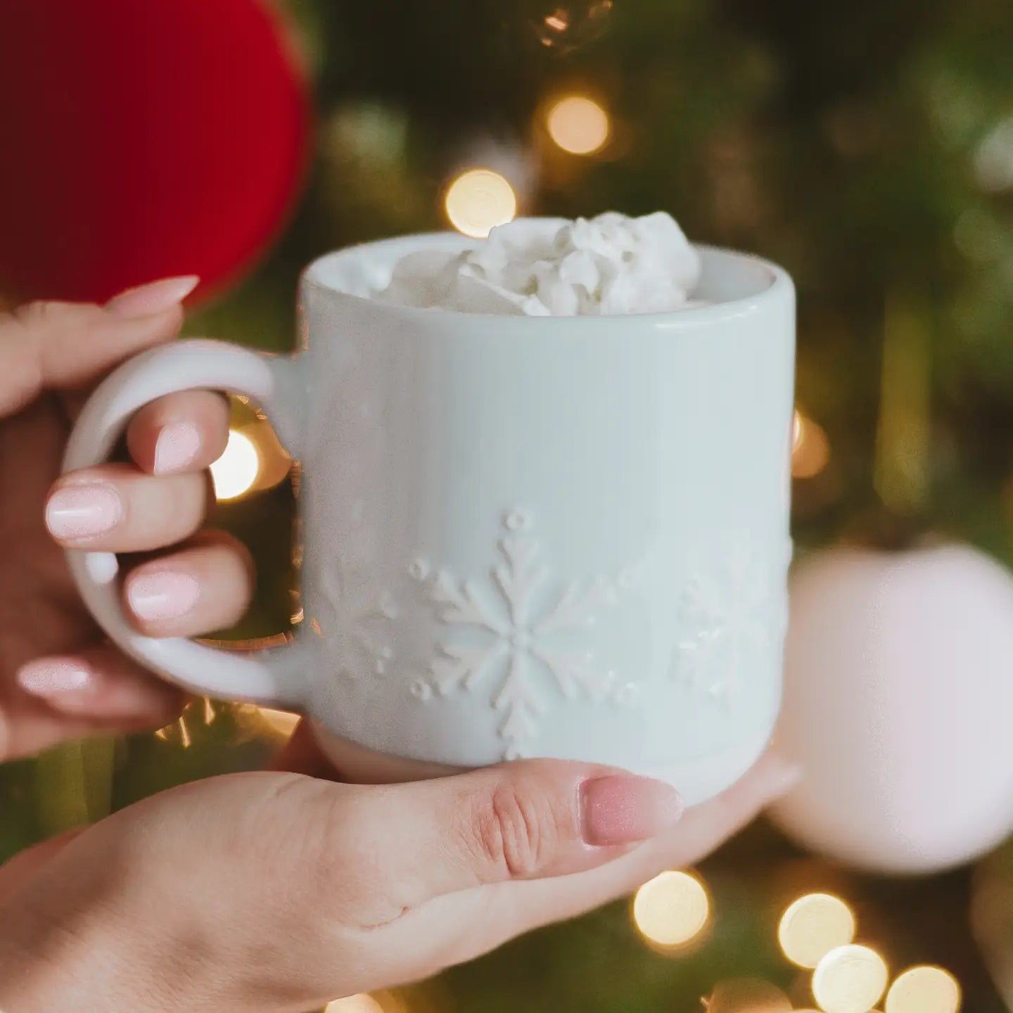 White mug with snowflake design held by a hand against a festive background with lights and decorations.