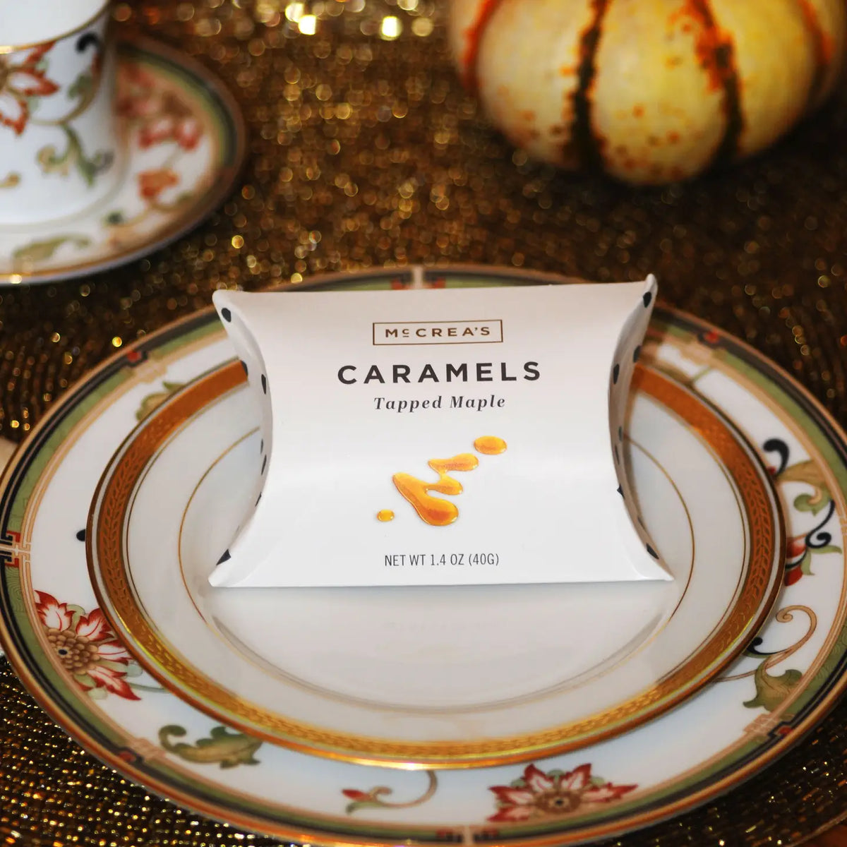 A white paper candy package labeled 'MSCREA'S CARAMELS Tapped Maple' sits centered on a decorative white plate with gold and floral detailing, set on a glittery gold tabletop with a small gourd in the background.