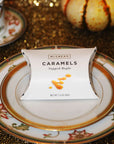 A white paper candy package labeled 'MSCREA'S CARAMELS Tapped Maple' sits centered on a decorative white plate with gold and floral detailing, set on a glittery gold tabletop with a small gourd in the background.