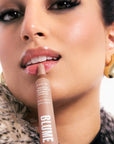 The image shows a close-up of a person applying "Blume Superbalm" to their glossy lips. The tube is a beige-pink shade with text indicating the flavor "Vanilla Cream" and mentions a "Tri-Peptide" formula. The individual has radiant, smooth skin with subtle makeup and is wearing statement gold earrings. A soft fur-textured coat is visible, adding to the luxurious feel of the scene. The person’s lips are glossy, hinting at the moisturizing and hydrating qualities of the balm.