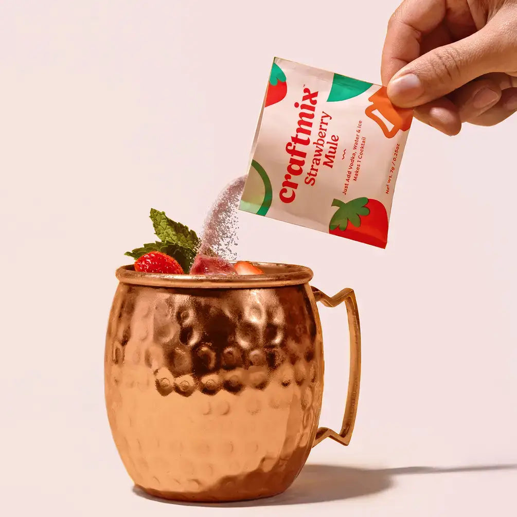 Hand pouring a Craftmix Strawberry Mule mix packet into a copper mug with strawberries and mint leaves.