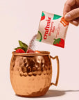 Hand pouring a Craftmix Strawberry Mule mix packet into a copper mug with strawberries and mint leaves.