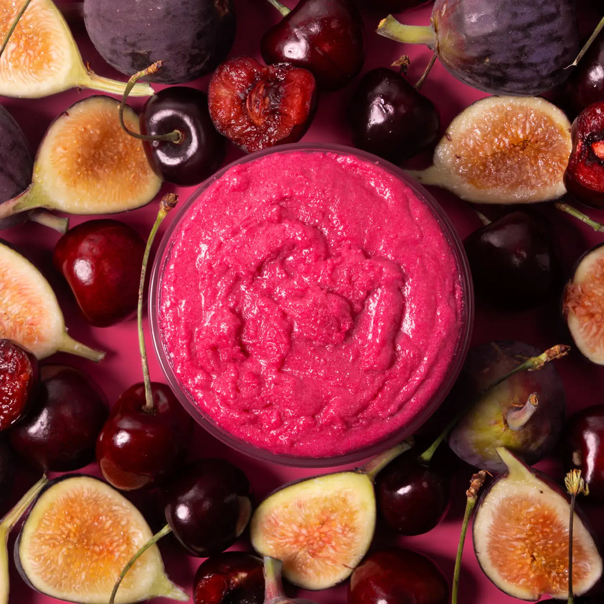 A round glass jar filled with bright pink berry spread, surrounded by fresh figs and cherries on a matching pink background.