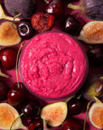 A round glass jar filled with bright pink berry spread, surrounded by fresh figs and cherries on a matching pink background.