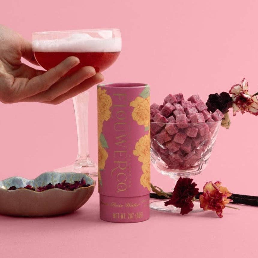 Hand holding a glass of red drink with a pink container labeled 'Flower Co.' on a pink background.