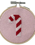 close-up of the same candy cane cross-stitch design within a wooden embroidery hoop. The candy cane is stitched with alternating red and white stripes on a pink background, creating a playful and festive look perfect for holiday decorations or gifts.