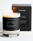A lit True Hue Santal soy wax blend candle in a clear glass jar with a black label, shown in front of its black product box on a clean white background.