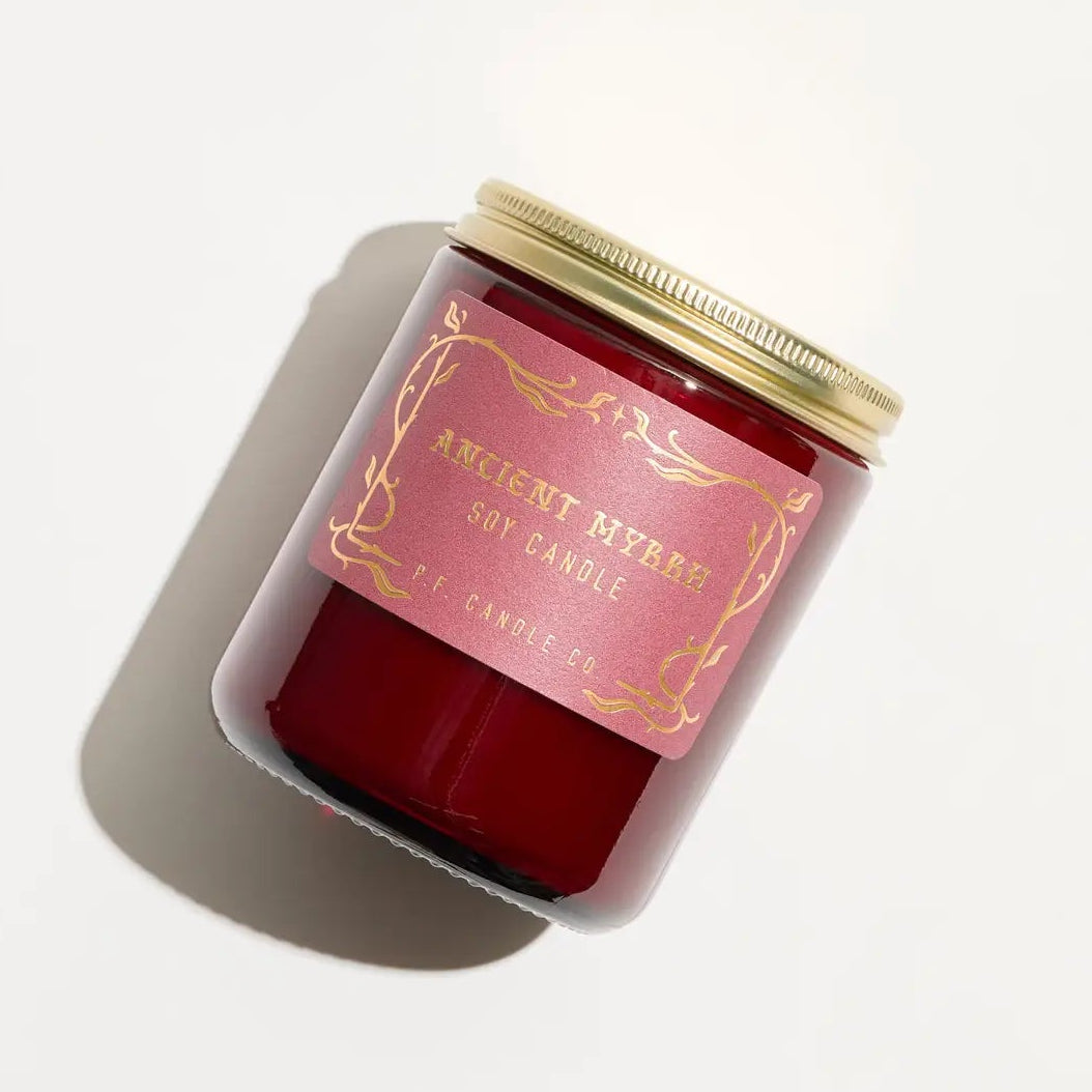 Red candle with gold top and gold writing that states "ancient myrth soy candle" 