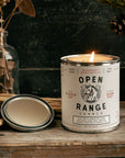 Candle in a can labeled 'Open Range Candle' on a wooden surface with decorative elements.