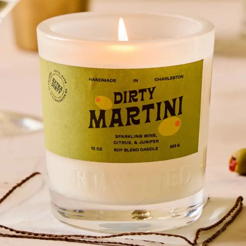 A candle labeled "Dirty Martini" with a green label, featuring notes of sparkling wine, citrus, and juniper. The candle sits on a coaster, lit and creating a cozy ambiance. In the background, there are martini glasses, one with an olive garnish, a gold cocktail shaker, and additional cocktail-making accessories, evoking a sophisticated and playful cocktail-inspired setting.