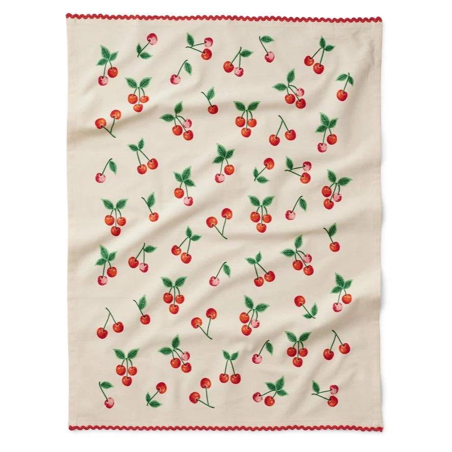 A tea towel laid flat, featuring a cream-colored background with a pattern of bright red cherries and green leaves scattered across. The edge is adorned with a red ric-rac trim for a playful, vintage-inspired look.