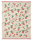 A tea towel laid flat, featuring a cream-colored background with a pattern of bright red cherries and green leaves scattered across. The edge is adorned with a red ric-rac trim for a playful, vintage-inspired look.