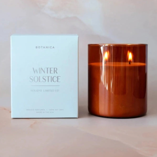 The image shows a candle product from the brand "Botanica" with the label "Winter Solstice" and "Holiday Limited Ed" on the packaging. The packaging is a simple, light-colored box with elegant, minimalist text, giving it a festive and serene feel. Next to the box is the candle, housed in a warm amber-colored glass container with two lit wicks, creating a cozy, inviting glow. The background features a soft, pastel marble or blurred surface, enhancing the warm and luxurious appearance of the product.