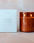 The image shows a candle product from the brand "Botanica" with the label "Winter Solstice" and "Holiday Limited Ed" on the packaging. The packaging is a simple, light-colored box with elegant, minimalist text, giving it a festive and serene feel. Next to the box is the candle, housed in a warm amber-colored glass container with two lit wicks, creating a cozy, inviting glow. The background features a soft, pastel marble or blurred surface, enhancing the warm and luxurious appearance of the product.